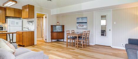 Een ruime woonkamer met houten vloer, een eettafel met stoelen, een keuken met houten kastjes en een grijze bank.