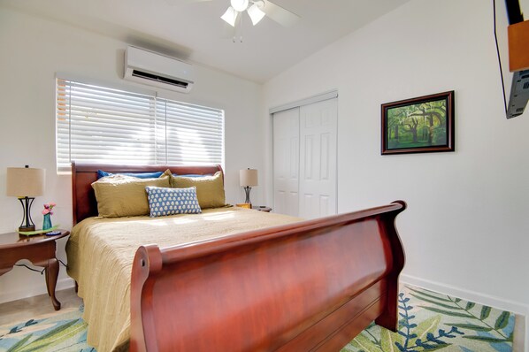 Un dormitorio con cama de madera, paredes blancas, ventilador de techo, unidad de aire acondicionado, un cuadro enmarcado y una ventana con persianas.