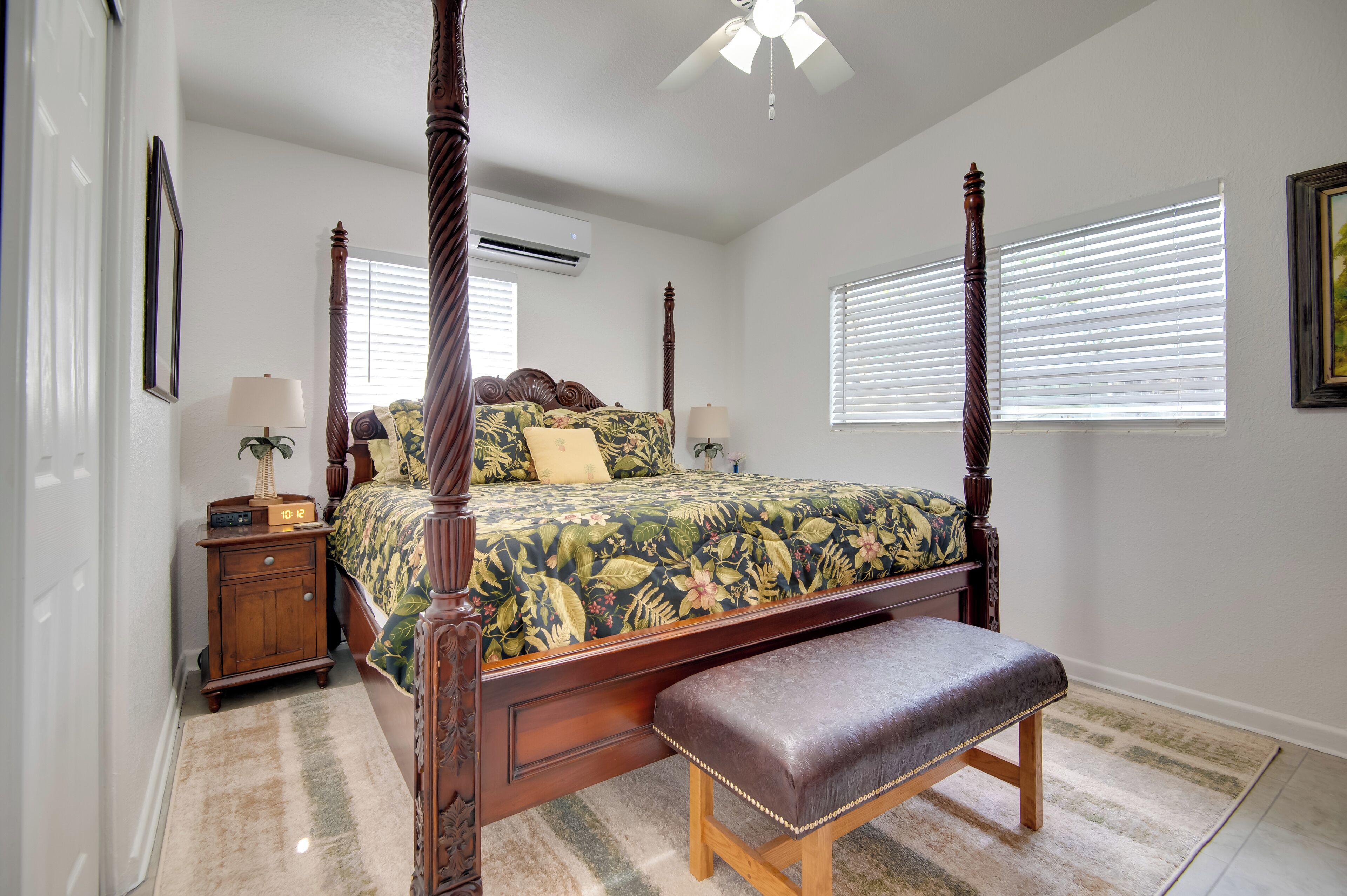 A four-poster bed with a floral bedspread, a wooden bench, and a nightstand with a lamp.