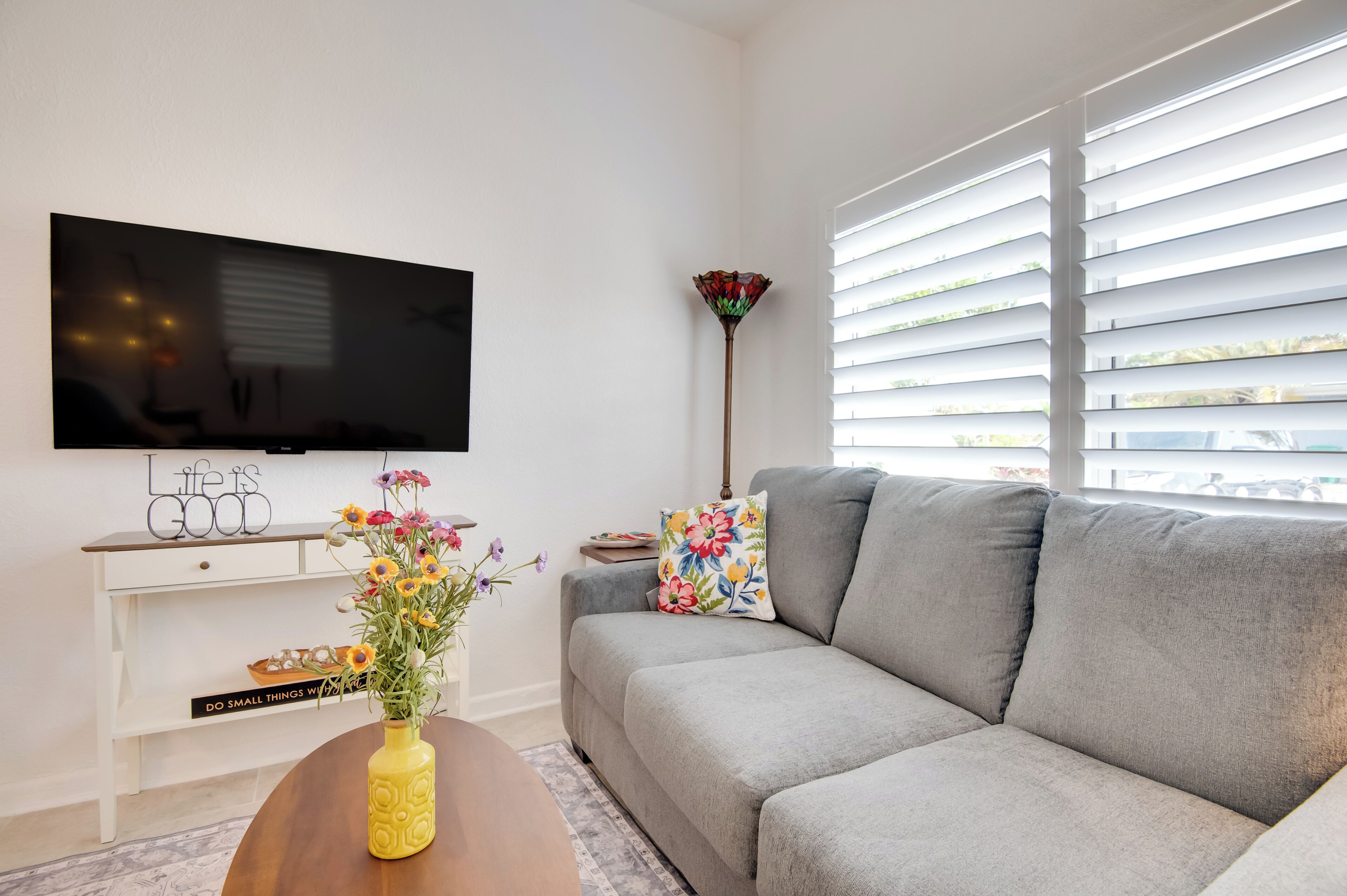 A modern living room with a grey sofa, a flat-screen TV mounted on the wall, a white console table with decorative items, and a colorful vase on a wooden coffee table.