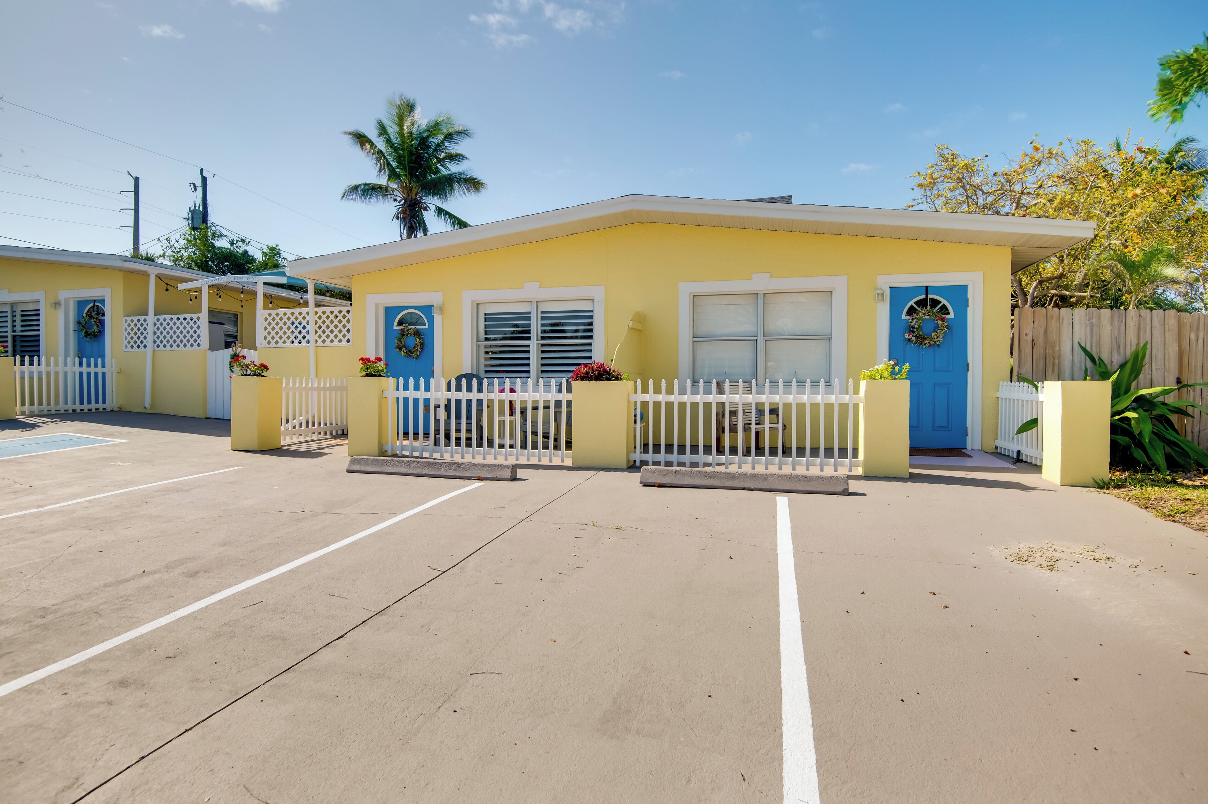 A row of yellow buildings with white fences and blue doors, each with a wreath and potted plants.