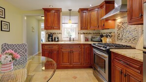 A kitchen with wooden cabinets, a stove, and a glass table with a vase of flowers. - 1 Mi to South Village Beach: Bbq-ready Retreat (West Dennis)