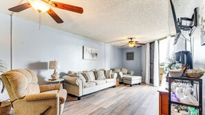 Una sala de estar con un sofá, un sillón y un ventilador de techo. Hay una mesita con una lámpara y una estantería con varios objetos.