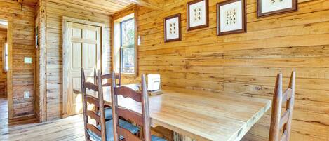 A wooden interior with a dining table and chairs, framed pictures on the wall, and a door leading outside.