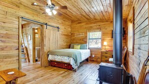 Camera da letto in una baita di legno, con un letto, pavimento in legno, ventola a soffitto, una finestra e un camino a legna.