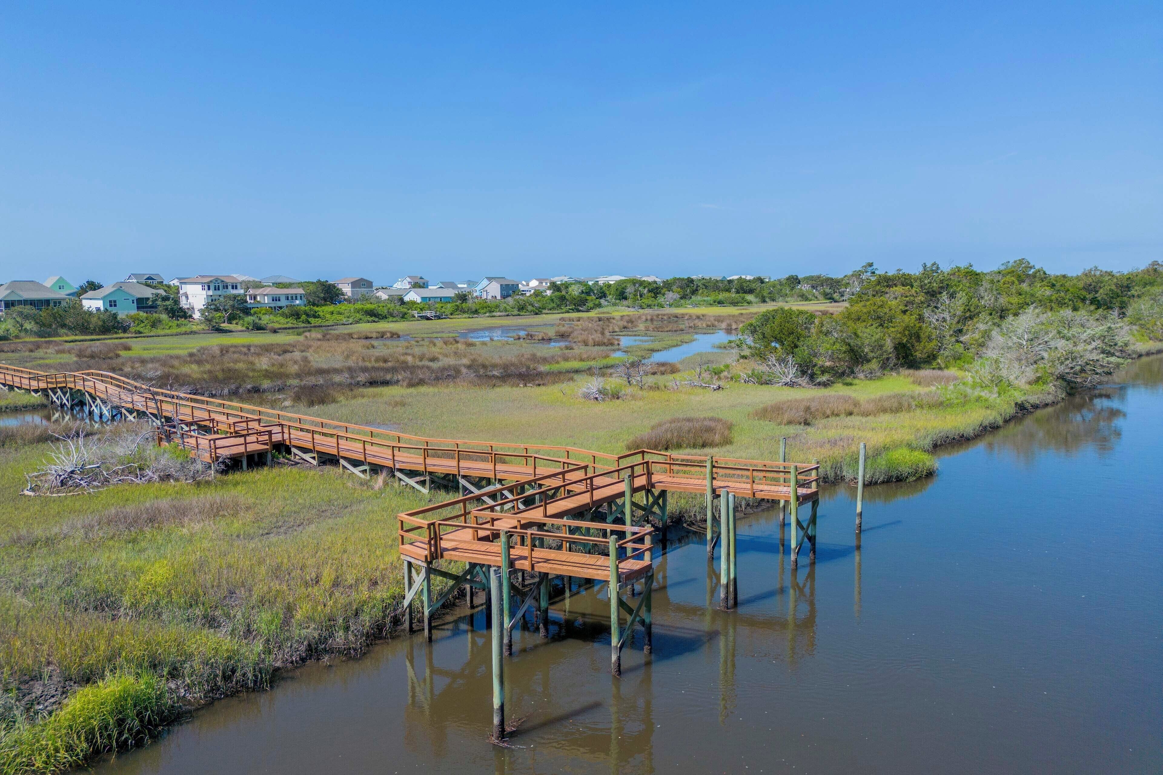 Un paseo marítimo de madera sobre una zona pantanosa con un río tranquilo, rodeado de vegetación y casas al fondo.