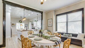 A dining area with a large oval table set for six, surrounded by wooden chairs. A kitchen is visible in the background, and there is a window with blinds on the right side.