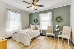 Un dormitorio con cama, dos sillas, ventilador de techo y una mesita de noche con un florero.