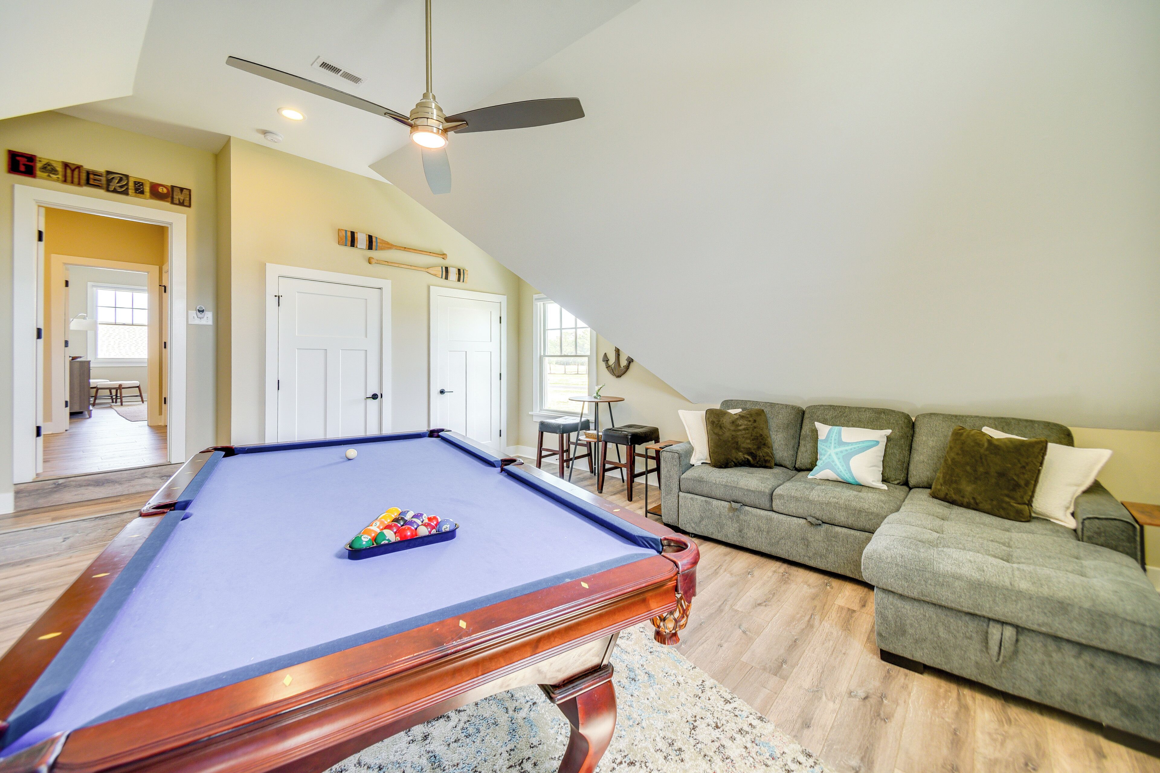 Une table de billard dans un salon, avec un ventilateur de plafond, un canapé gris avec des coussins et un plancher en bois.
