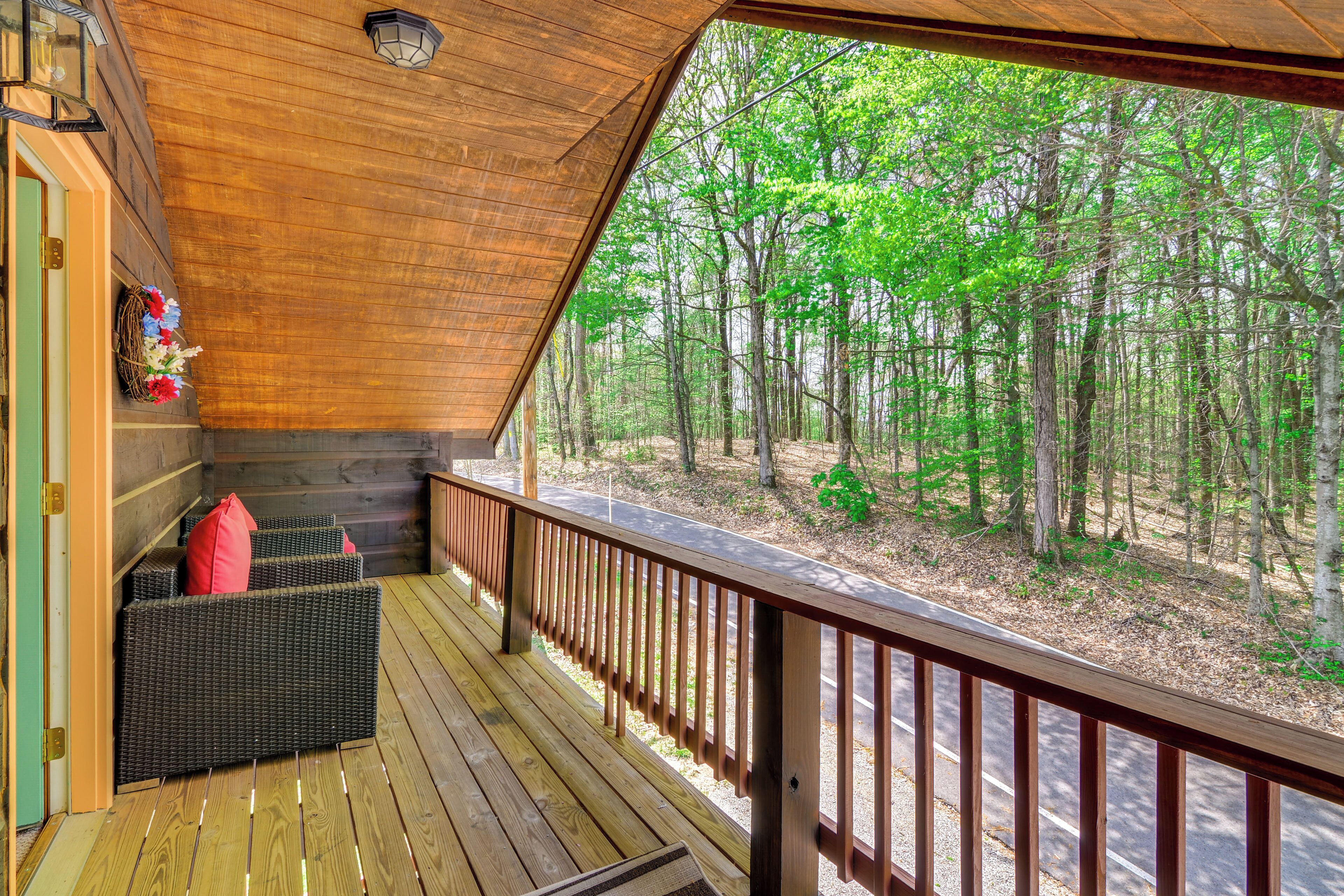 A wooden deck with a wicker sofa and red cushions, overlooking a forested area.