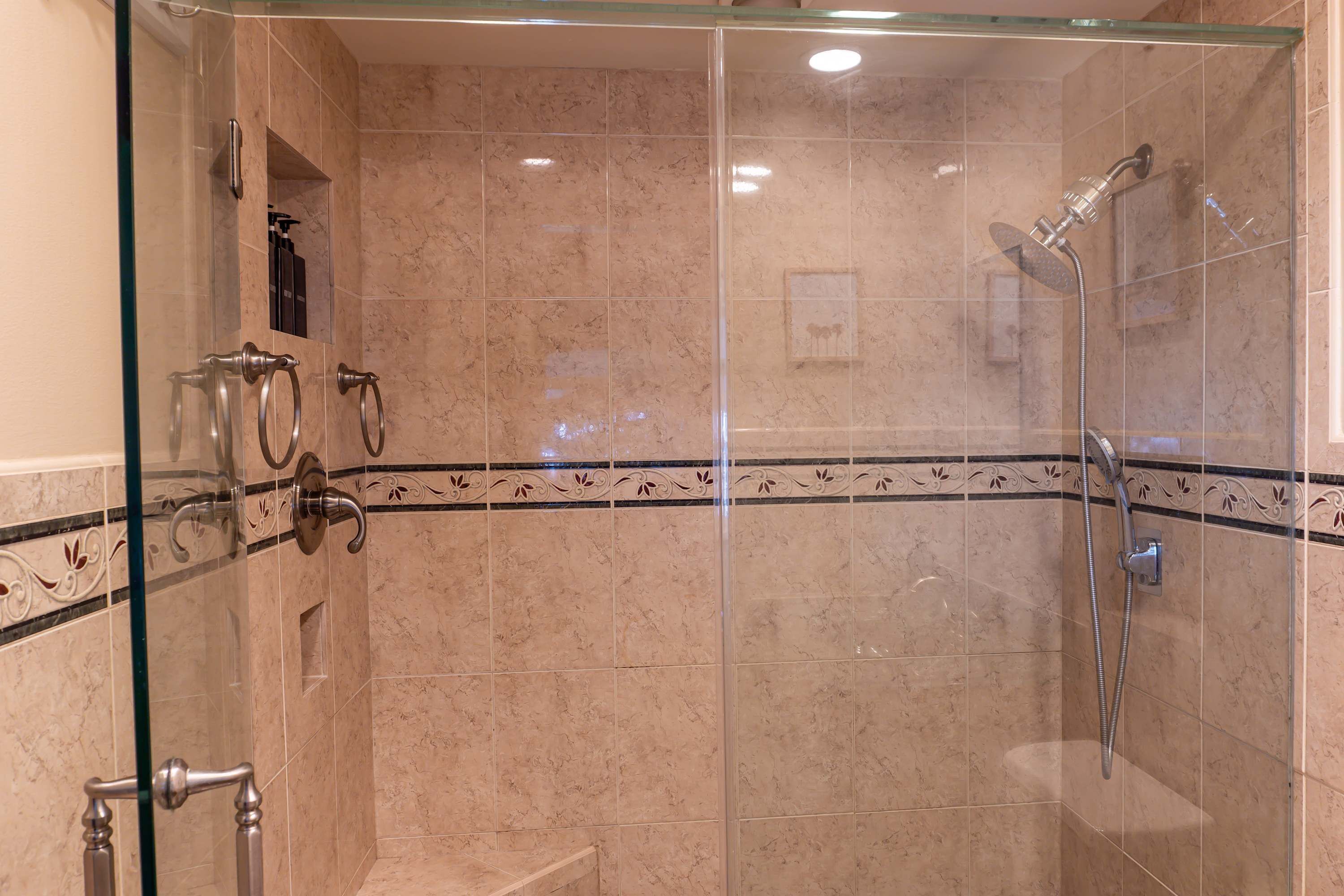 A glass-enclosed shower area with a marble-tiled wall, a showerhead, and a handrail.