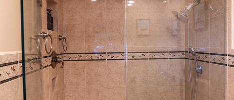 A glass-enclosed shower area with a marble-tiled wall, a showerhead, and a handrail.