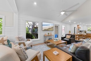 A spacious living room with a large window, a ceiling fan, and a dining area with wooden furniture. - Kiawah Exclusives | 459 Oceanwoods Cottage (Kiawah Island)