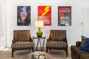 Uma sala de estar com duas poltronas listradas, uma mesa de centro e três cartazes de filmes na parede.