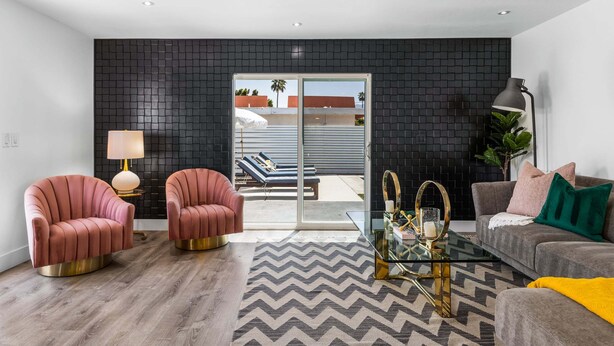 Ein modernes Wohnzimmer mit schwarzer Wand aus Fliesen, zwei rosafarbenen Sesseln, einem Glastisch, einem grauen Sofa mit bunten Kissen und Blick auf einen Balkon mit Pool.