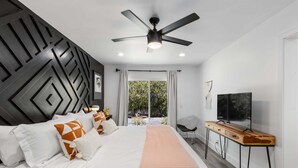 Ein modernes Schlafzimmer mit Bett, Deckenventilator, Fernseher und Blick auf Grünfläche.