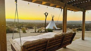 A wooden swing chair with a white cushion, overlooking a desert landscape with a white teepee and hanging lights. - Navigate Stays retreat W Hot Tub Bobs Homestead (Joshua Tree)