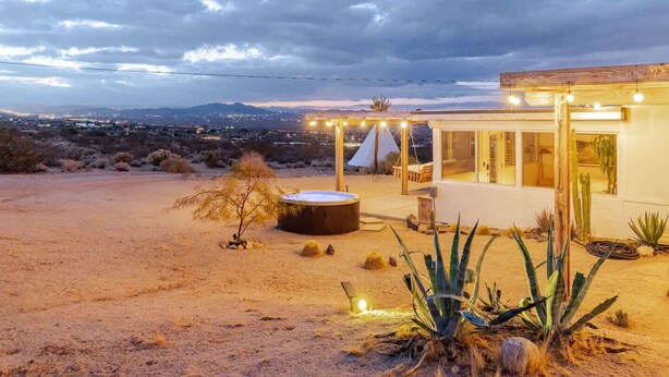 A desert landscape with a modern house featuring a hot tub, outdoor seating, and cacti. - Navigate Stays retreat W Hot Tub Bobs Homestead (Joshua Tree)