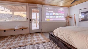 A bedroom with a bed, a projection screen displaying apps and games, a wooden ceiling fan, and a view through a door. - Navigate Stays retreat W Hot Tub Bobs Homestead (Joshua Tree)