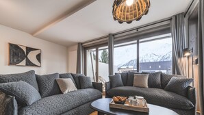 Un salon moderne avec deux canapés gris foncé, une table basse sur laquelle est posée une assiette de pâtisseries, et une grande fenêtre offrant une vue sur des montagnes enneigées.