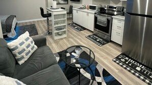 Una sala de estar moderna con un sofá gris, una mesita de centro y un televisor. La cocina cuenta con gabinetes blancos, un microondas y un refrigerador.