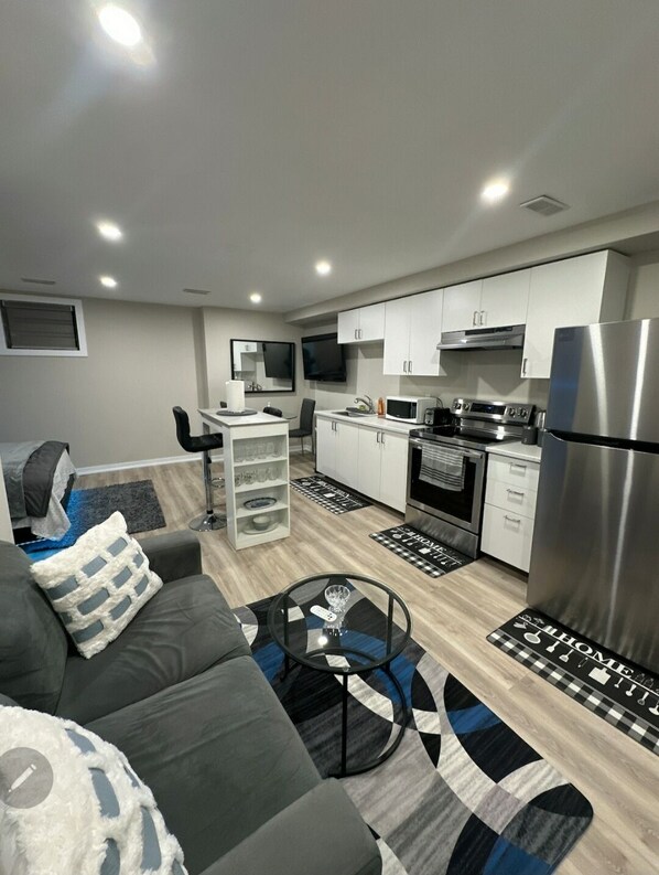 Una sala de estar moderna con un sofá gris, una mesita de centro y un televisor. La cocina cuenta con gabinetes blancos, un microondas y un refrigerador.