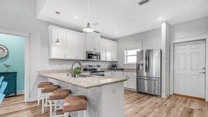 A modern kitchen with white cabinets, a central island with bar stools, stainless steel appliances, and pendant lighting. - Gulf View Beach Retreat with Hot Tub (Gulf Shores)