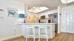 A modern kitchen with white cabinetry, a central island with bar stools, a microwave, a refrigerator, and a ceiling light fixture. - 7th Floor Beachfront Condo w/ Beachside Bar & Resort Pool in Destin! 🐚💙🌴 (Destin)