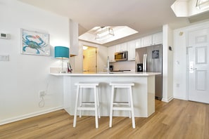 A modern kitchen with white cabinetry, a central island with bar stools, a microwave, a refrigerator, and a ceiling light fixture.