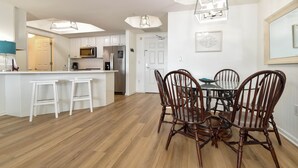 A modern kitchen with white cabinets, a central island with stools, and a dining area with wooden chairs. - 7th Floor Beachfront Condo w/ Beachside Bar & Resort Pool in Destin! 🐚💙🌴 (Destin)