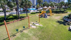 A playground with swings, a slide, and a climbing structure, surrounded by palm trees and a pool.