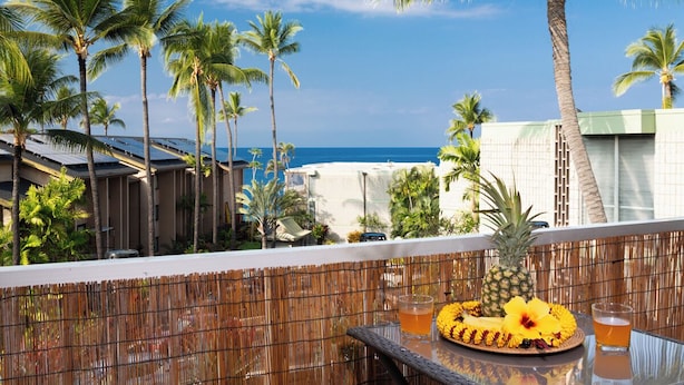 A balcony with a table set for a meal, overlooking a coastal area with palm trees and a building with a green roof.
