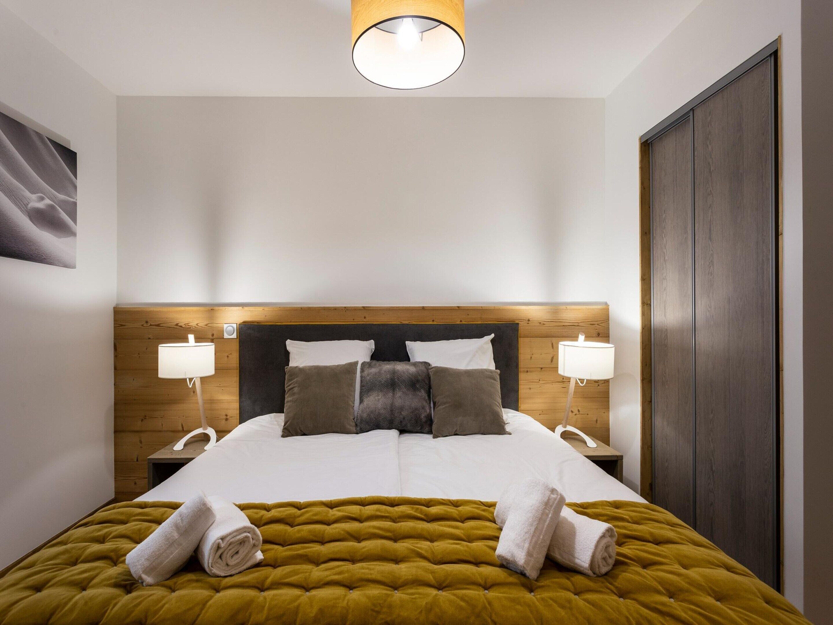A neatly made bed with a mustard-colored duvet, two white towels, and a wooden headboard.