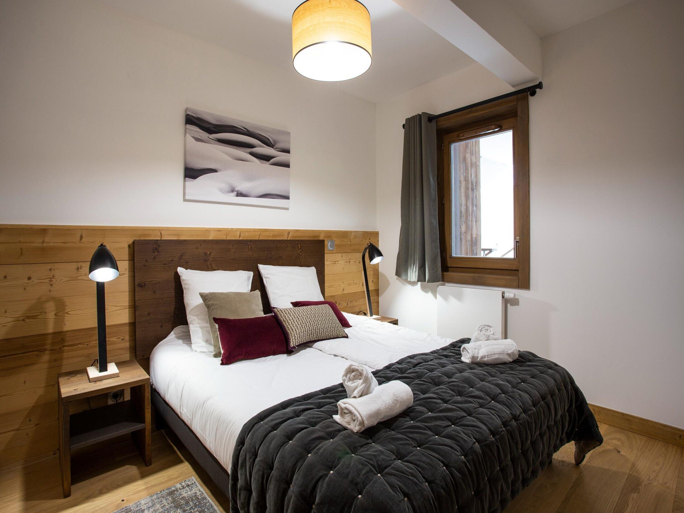 Een slaapkamer met een houten hoofdbord, een bed met witte lakens en meerdere kussens, een zwart geborduurde spreideken, twee bedlampjes, een raam met gordijnen en een schilderij aan de muur.