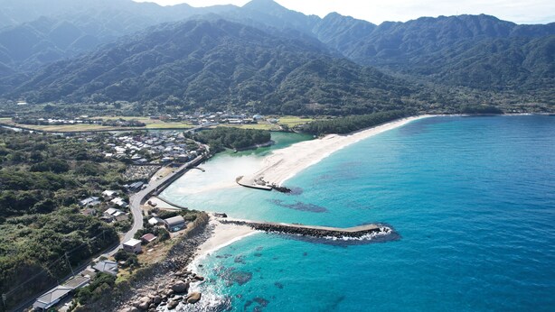 An aerial view of a coastal area with a beach, a rocky breakwater, and a residential area with houses and a road. - Nagata Sea Time - Private Retreat in Yakushima, Near the Sea Turtle Beach (熊毛郡屋久島町永田)