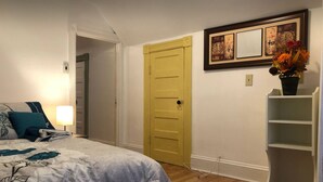 A bedroom with a bed, a nightstand, a lamp, a yellow door, and a wall-mounted shelf with a vase of flowers. - Cozy Modern City Escape (Schenectady)
