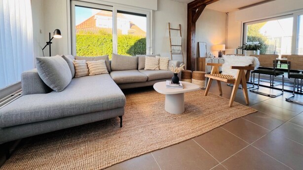 A modern living room with a grey sectional sofa, a round coffee table, and a dining area with a wooden table and chairs. - Holiday Home in Wenduine Near Zeedijk (De Haan)