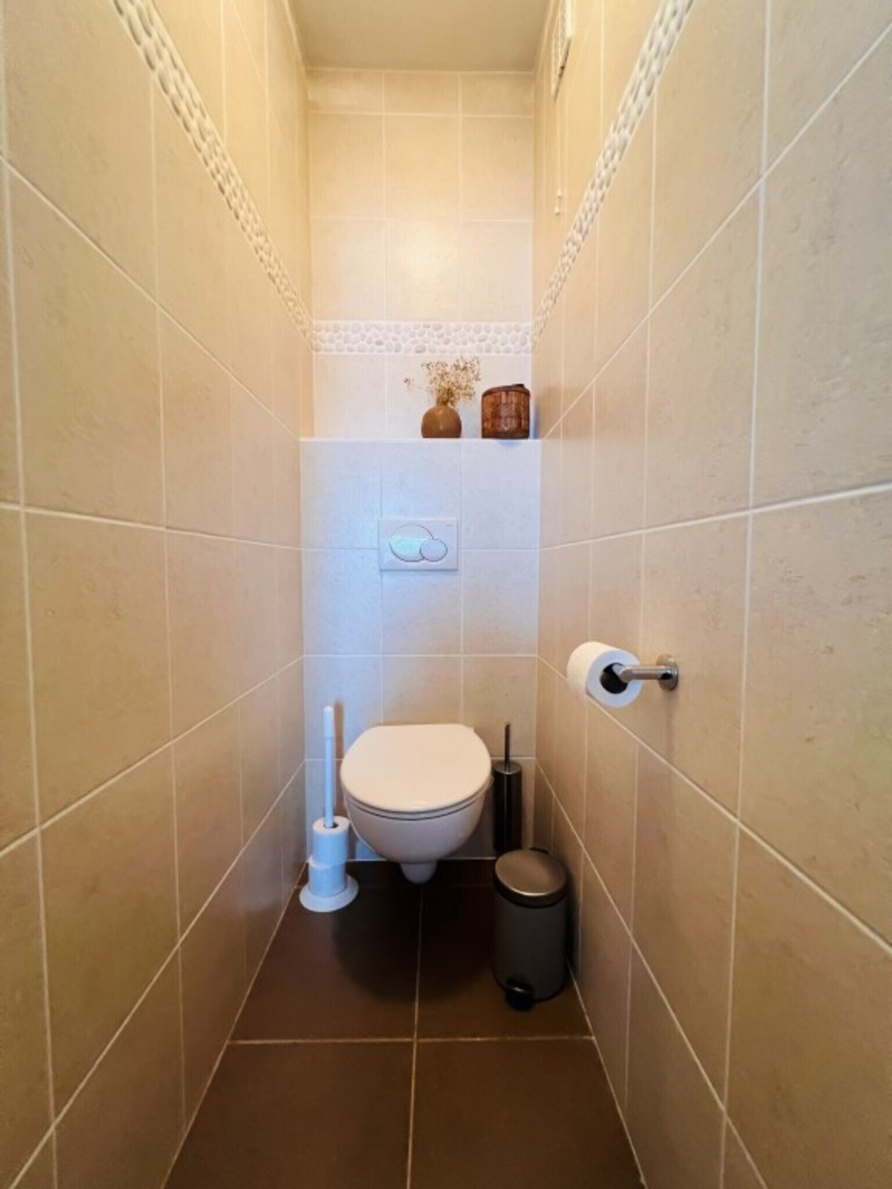 A small, narrow bathroom with beige tiles, a toilet, a bidet, a trash bin, and a toilet paper holder.