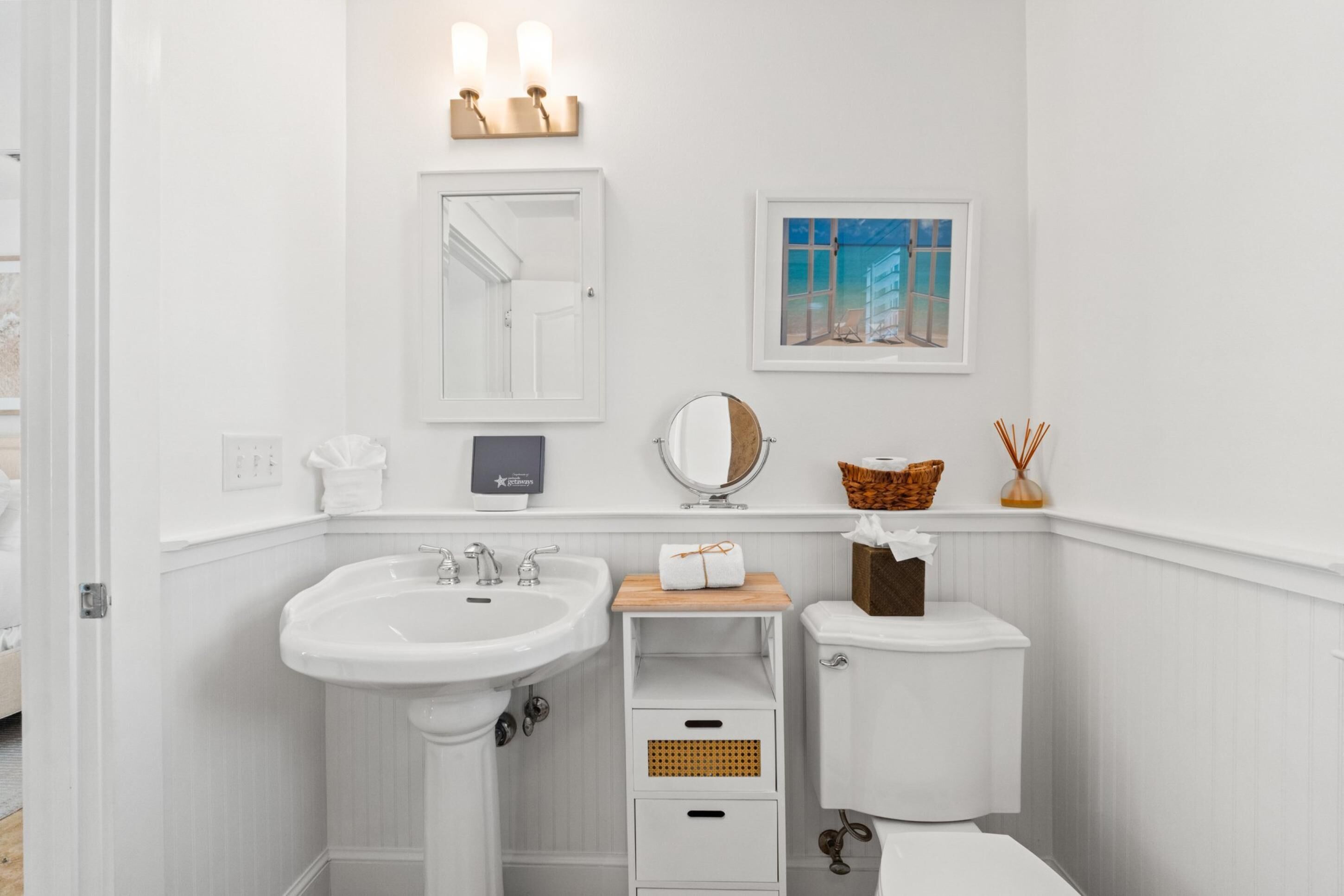 A white bathroom with a pedestal sink, a toilet, a mirror, and a framed picture on the wall.