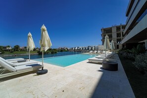 A swimming pool with lounge chairs and umbrellas, surrounded by a modern building and a view of the water.