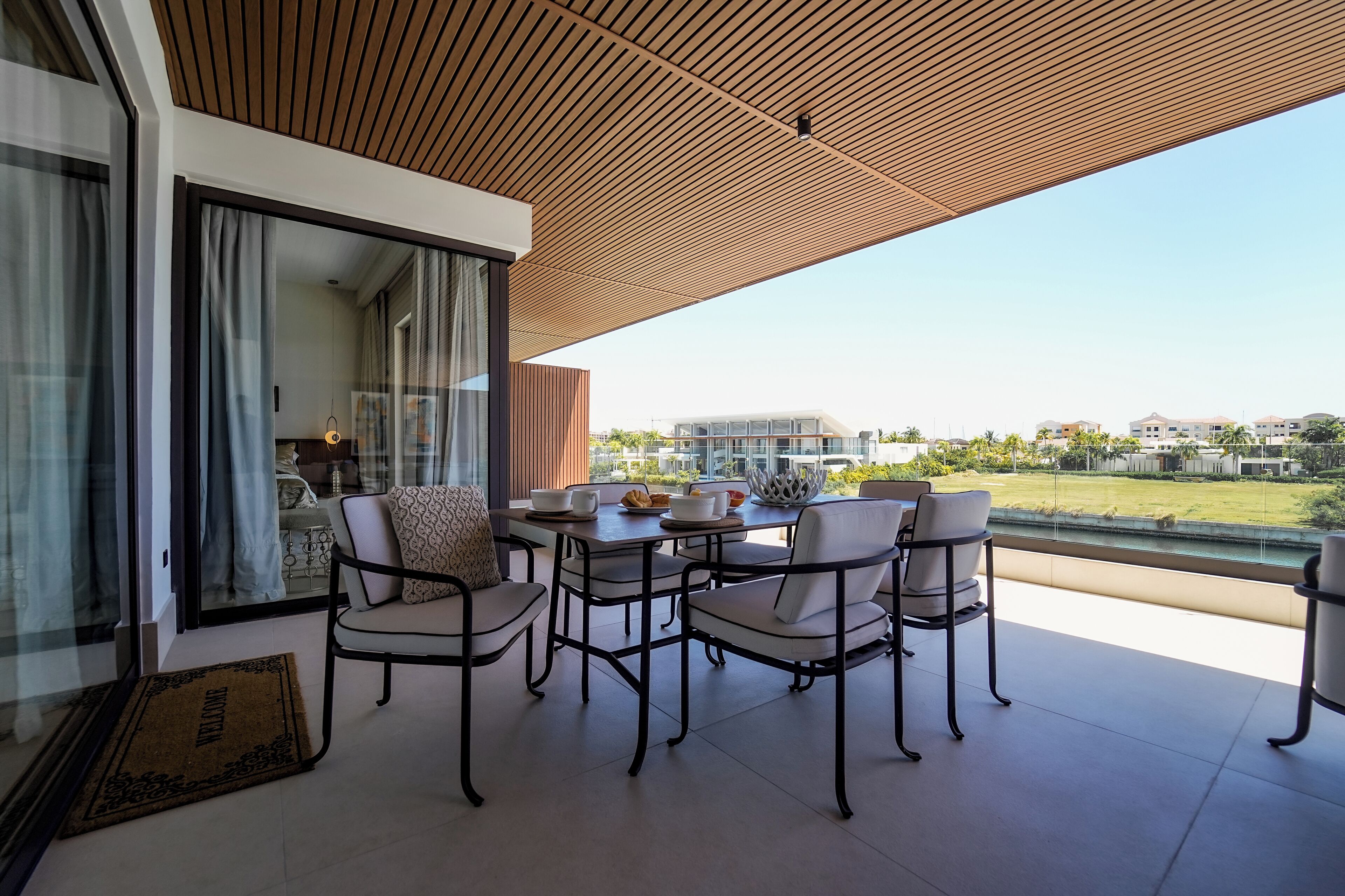 A modern outdoor dining area with a wooden ceiling, a glass door leading to a room, and a view of a residential area.
