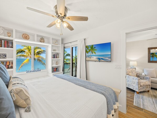 A bedroom with a bed, a ceiling fan, a bookshelf, a TV, and a view of the beach through a sliding door. - Seaside 306 | Stunning Beachfront, Newly Updated + Resort Amenities (Fort Myers Beach)