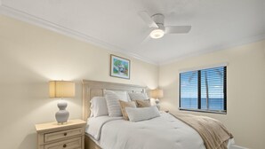 A bedroom with a bed, nightstands, a ceiling fan, a window with blinds, and a painting on the wall. - Seaside 106 | Beachfront Renovated Condo with Gulf Views, Pool & Coastal Charm (Fort Myers Beach)