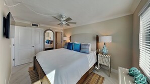 A bedroom with a bed, a ceiling fan, a nightstand with a lamp, a mirror, and a window with blinds.