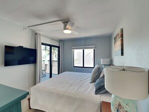 Un dormitorio con cama, ventilador de techo, televisor, ventana con vista a palmeras y un balcón.