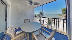 Ein Balkon mit rundem Tisch und vier Korbstühlen, der Blick auf einen Strand mit Palmen und den Ozean gewährt.