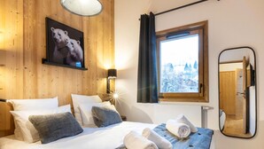 Une chambre confortable avec une tête de lit en bois, une grande fenêtre donnant sur des arbres couverts de neige, un lit avec du linge de lit blanc et des oreillers gris, et une lampe de chevet.