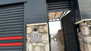 Entrada a una villa con muros de piedra y contraventanas de metal, un letrero que indica 'Villa Alen', y vista al mar de fondo.