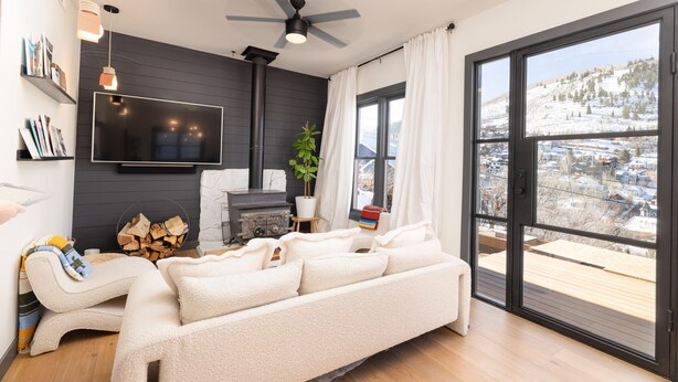 A modern living room with a sofa, a TV mounted on the wall, a ceiling fan, and a large window with a view of a snowy landscape.