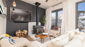A cozy living room with a fireplace, a TV mounted on the wall, a comfortable sofa, and a view of a snowy landscape through large windows.